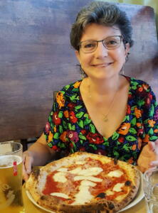Porträt von Frau Nolli-Meyer. Vor ihr steht auf einem Tisch eine großen Pizza und ein Getränk.