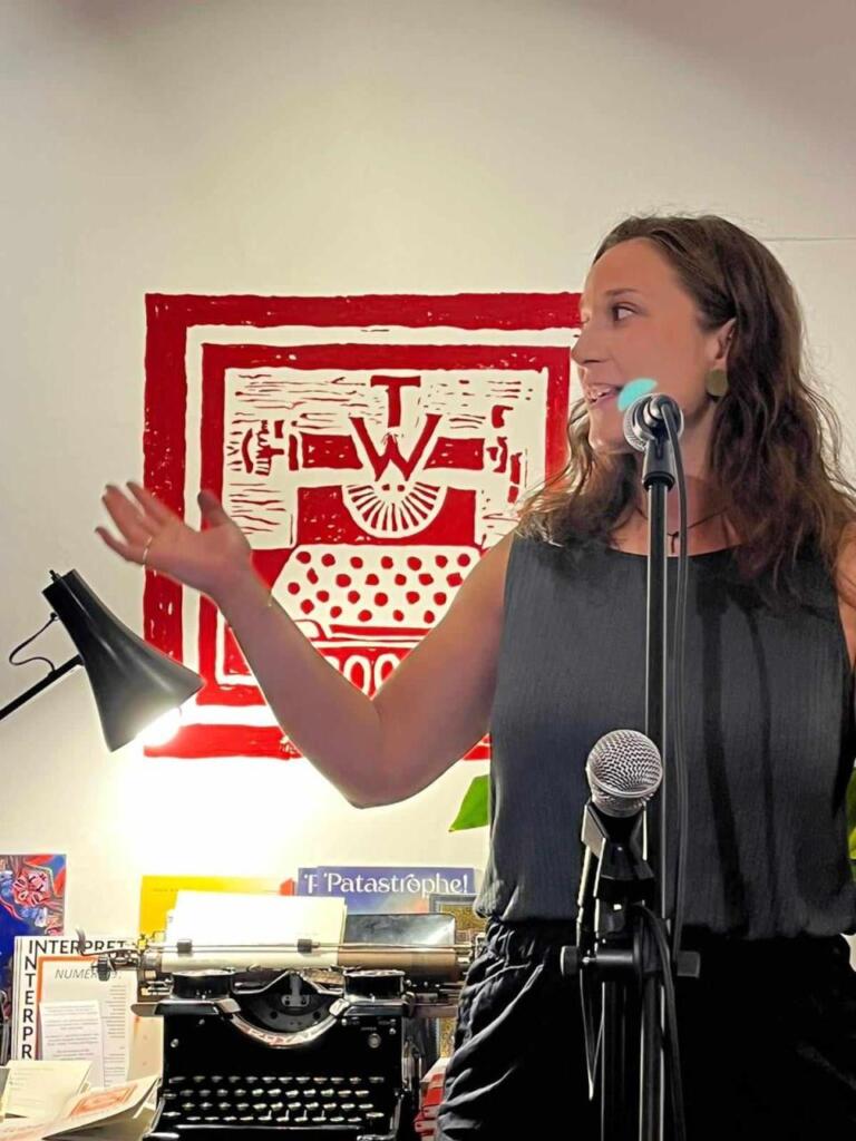 Eine Frau spricht in ein Mikrofon und zeigt mit der Hand auf die rechte Seite. Im Hintergrund eine Schreibmaschine und eine Lampe.