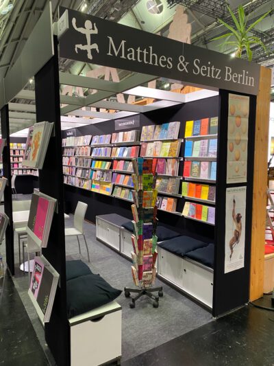 Blick in einen Bereich bei einer Messe. Über dem Eingang ein Schild „Matthes & Seitz Berlin“. Dahinter Sitzmöglichkeiten und ein Bücherregal.